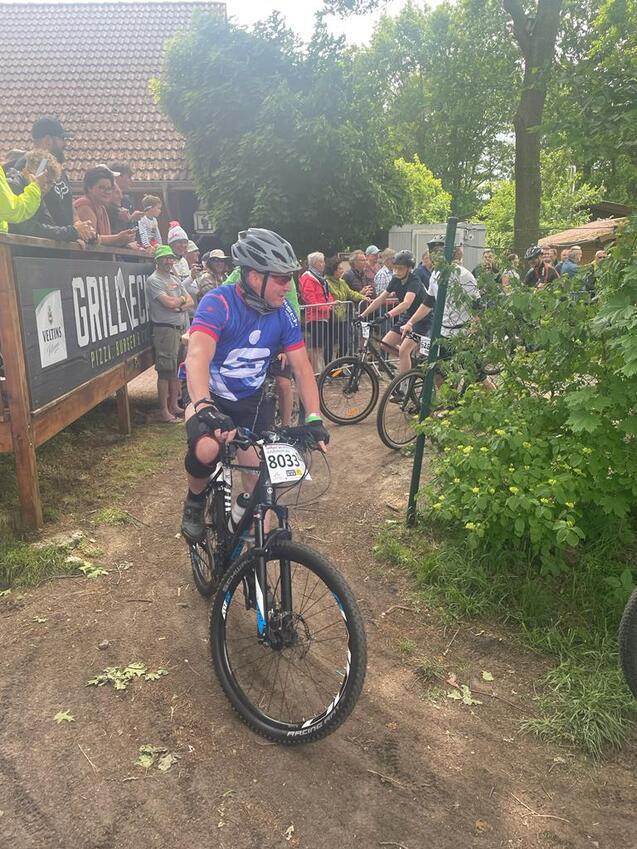 Mountainbiker in blauem Trikot fährt an Zuschauern vorbei, umgeben von Bäumen und einer Zielgeraden.