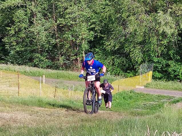 Zwei Mountainbiker fahren einen hügeligen Weg, umgeben von Bäumen und einem gelben Absperrzaun.