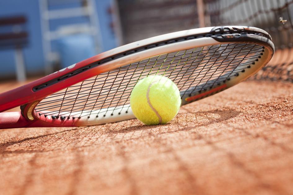 Tennisball liegt auf roter Sandplatz neben einem Tischtennisschläger mit Schlägerbespannung im Vordergrund.