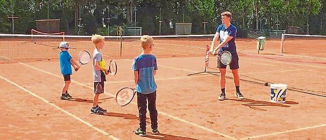 Trainer gibt vier Kindern auf einem Tennisplatz Anweisungen zum Tennisspielen, umgeben von Tennisnetzen.