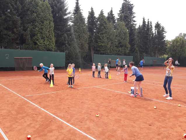Tennis-Training mit Kindern auf einem Sandplatz, betreut von zwei Erwachsenen, umgeben von Bäumen und Markierungen.