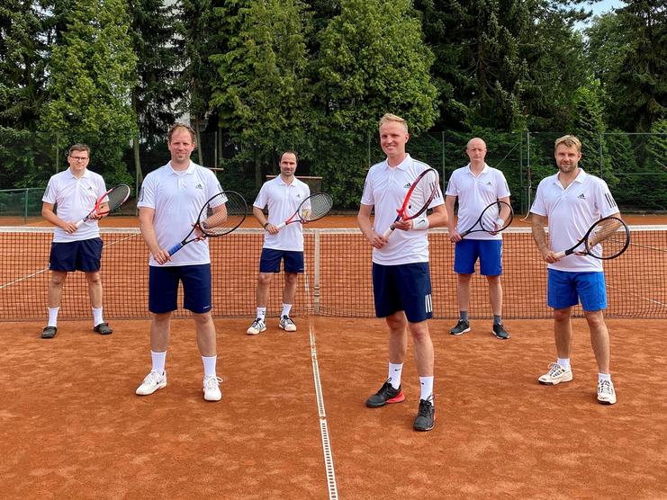 Sechs Männer in weißen Shirts und kurzen Hosen stehen auf einem Tennisplatz und halten Tennisschläger in der Hand.