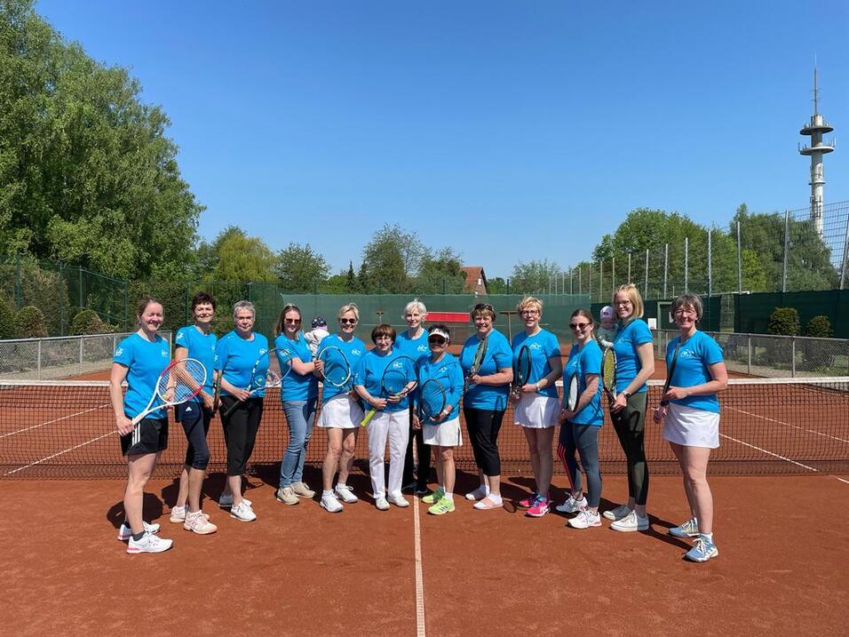 Gruppe von 14 Frauen in blauen T-Shirts steht auf einem Tennisplatz, bereit für ein Match bei sonnigem Wetter.