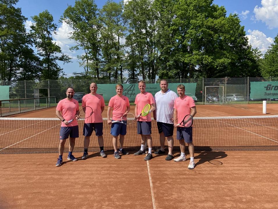 Sechs Männer in pinken Trikots stehen auf einem Tennisplatz, halten Schläger und lächeln vor grünen Bäumen.