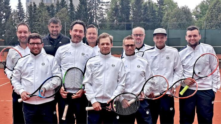 Eine Gruppe von zehn Tennisspielern in weißen Jacken steht auf einem Sandplatz, jeder hält einen Tennisschläger.
