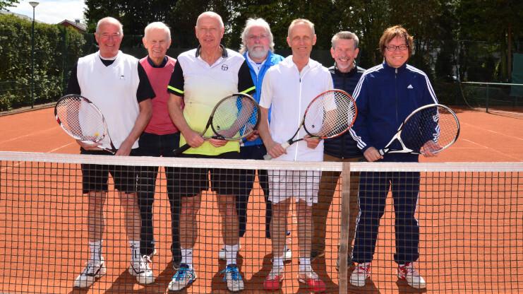 Gruppe von sieben älteren Männern mit Tennisschlägern auf einem Tennisplatz, lächelnd vor dem Netz stehend.