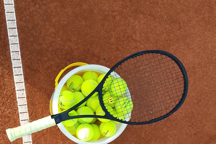 Tennisschläger liegt über einem Eimer mit leuchtend gelben Tennisbällen auf rotem Sandplatz.