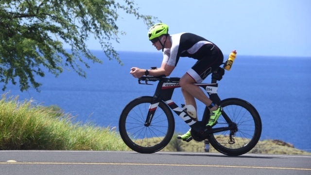 Sportler in Fahrradmontur fährt auf einer Straße mit Meerblick, Trinkflasche am Rahmen befestigt, sonniger Tag.