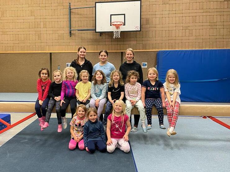Gruppe von Kindern und Betreuern in Sportkleidung, sitzend auf einer Turnbank in einer Sporthalle.