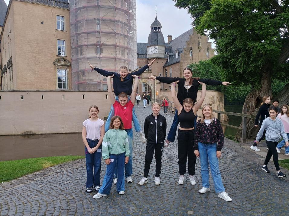 Gruppe von acht Jugendlichen posiert auf einem gepflasterten Weg vor einem historischen Gebäude mit einer Baustelle.