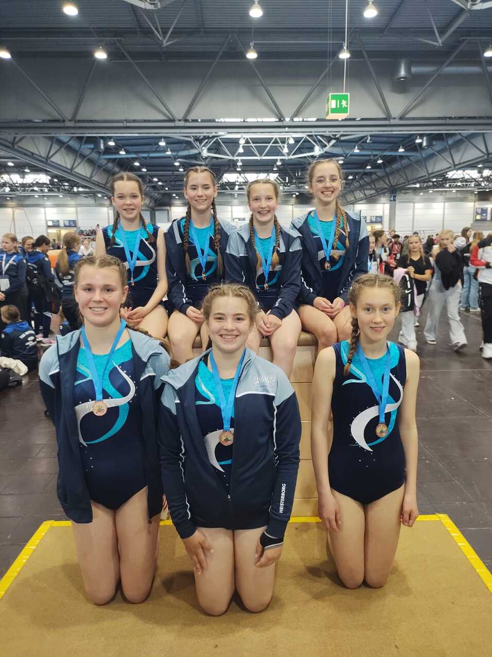 Gruppe junger Turnerinnen mit Medaillen posiert lächelnd auf einer Bühne in einer Sporthalle.