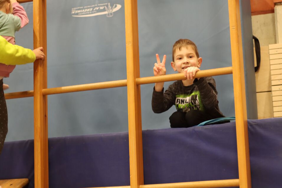 Junge sitzt auf einer Turnbank und zeigt ein Peace-Zeichen, während er an einem Holzgeländer festhält.