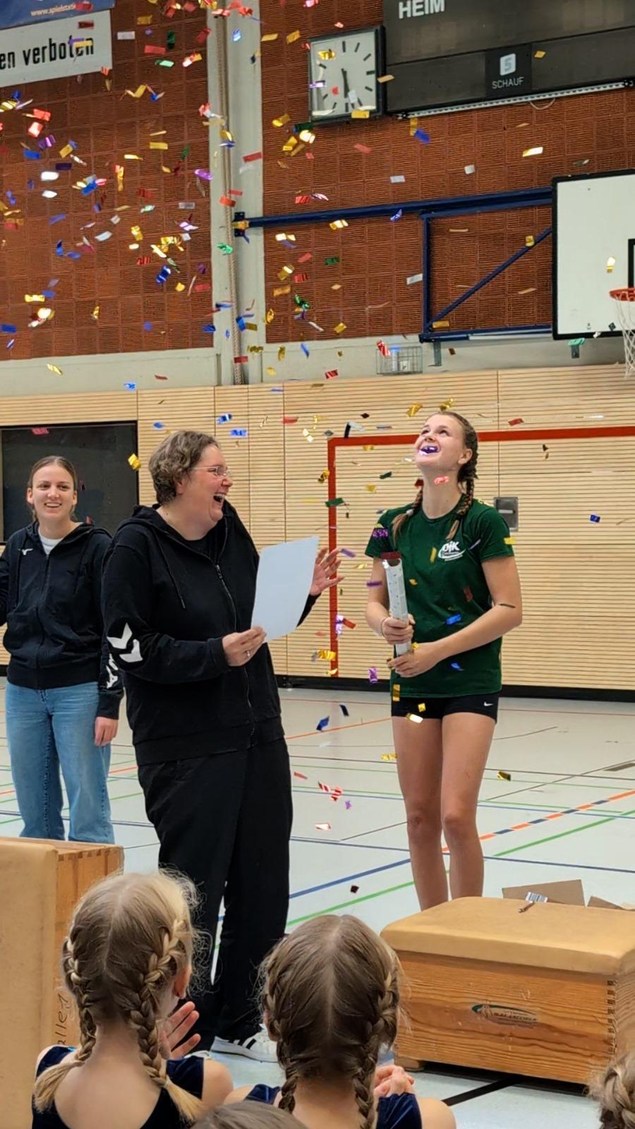 Lächelnde Frauen feiern mit Konfetti, eine Sportlerin hält einen Pokal und ein Zertifikat in einer Sporthalle.