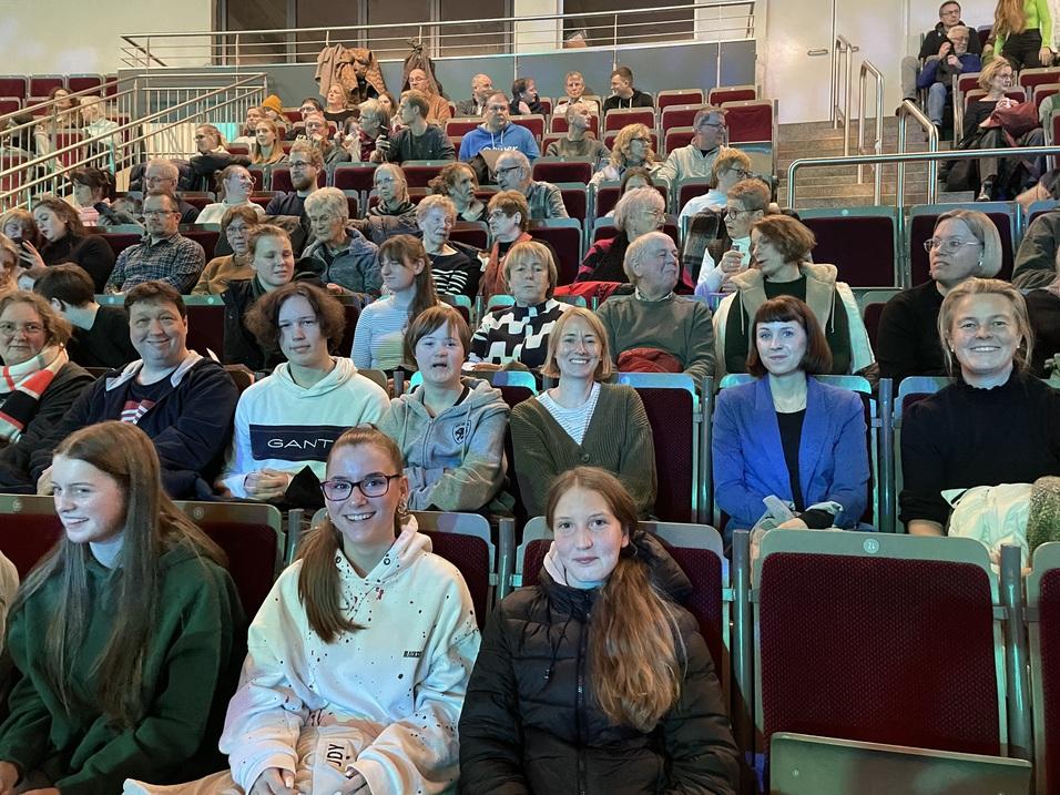 Gruppe von Menschen unterschiedlichen Alters sitzt in einer Aufführung, blickt zur Bühne mit bunten Theaterstühlen im Hintergrund.