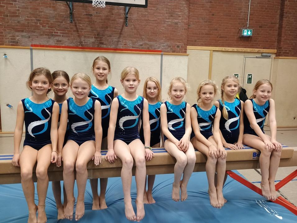 Gruppe von neun jungen Turnerinnen in blauen Bodenturnanzügen auf einem Schwebebalken in einer Turnhalle.