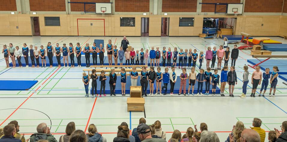 Gymnastikveranstaltung mit Gruppen von Kindern in Sportkleidung auf einer Matte, Zuschauer sitzen im Vordergrund.