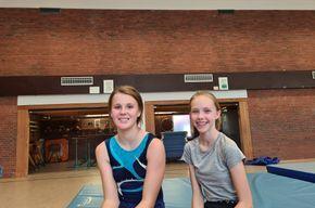 Zwei Mädchen lächeln auf blauen Turnmatten in einer Sporthalle mit Ziegelwänden.