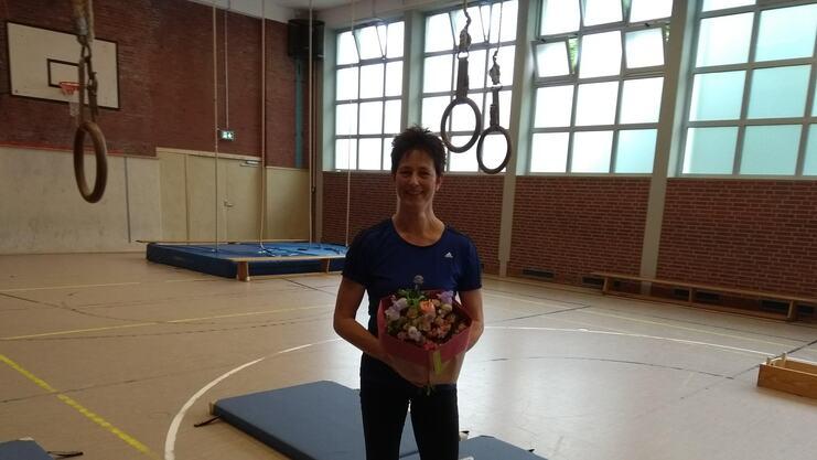 Frau mit einem Blumenstrauß steht in einer Turnhalle, im Hintergrund sind Turngeräte sichtbar.