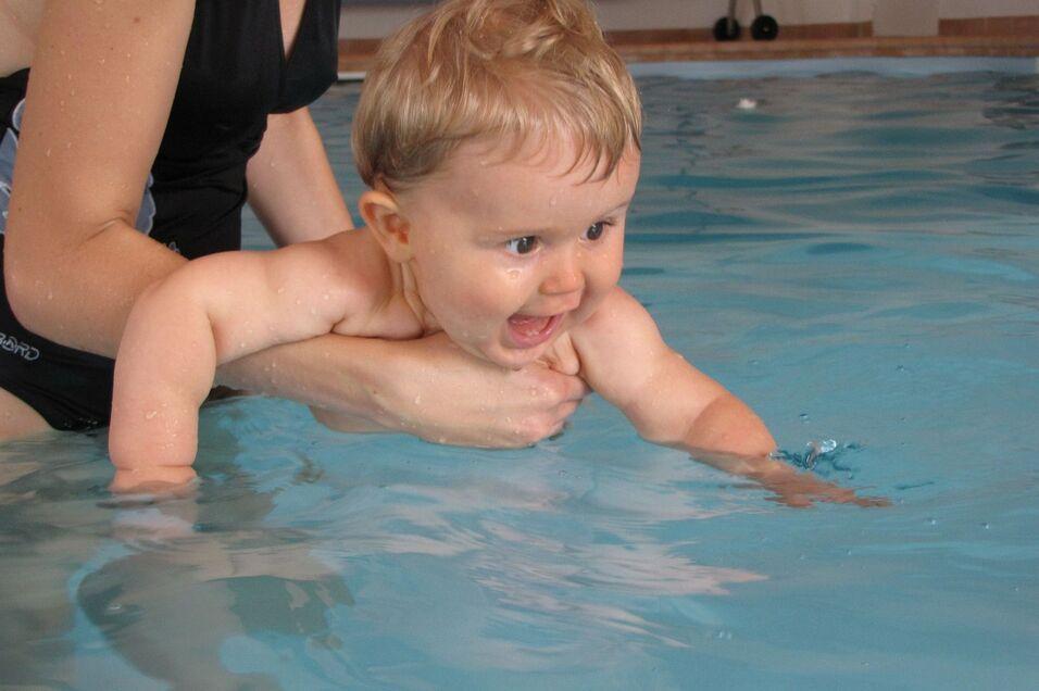 Ein Kleinkind wird von einer Erwachsenen im Wasser gehalten, fr&ouml;hlich mit Wasser spritzend.