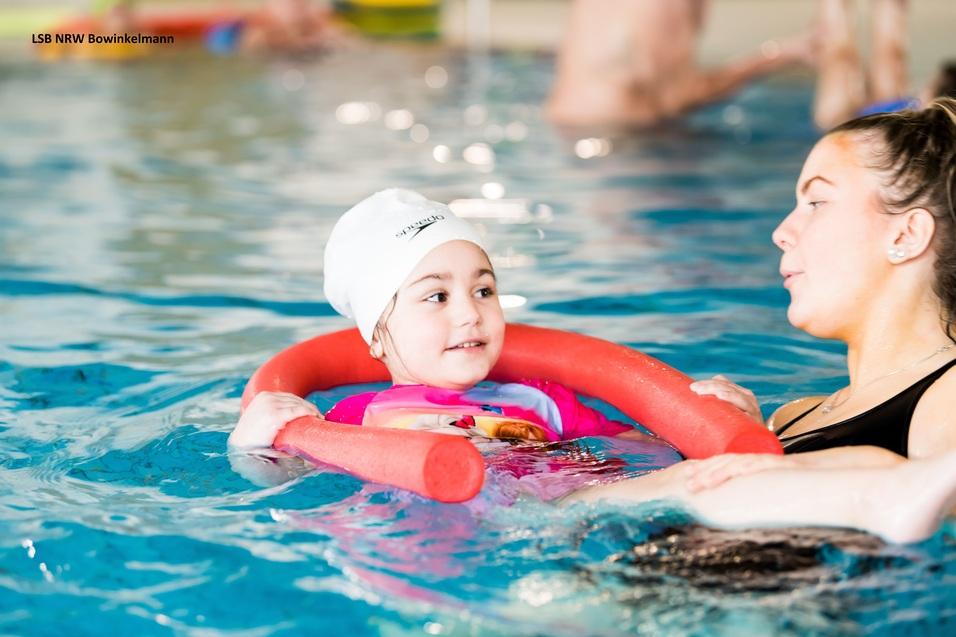 Ein kleines Mädchen mit einer Schwimmhilfe und Badekappe übt mit einer Trainerin im Schwimmbecken.