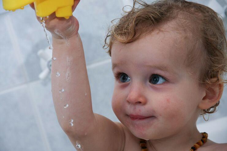 Kleines Kind mit lockigem Haar hält einen gelben Wasserspielzeug und spritzt Wasser in einer Badezimmerumgebung.