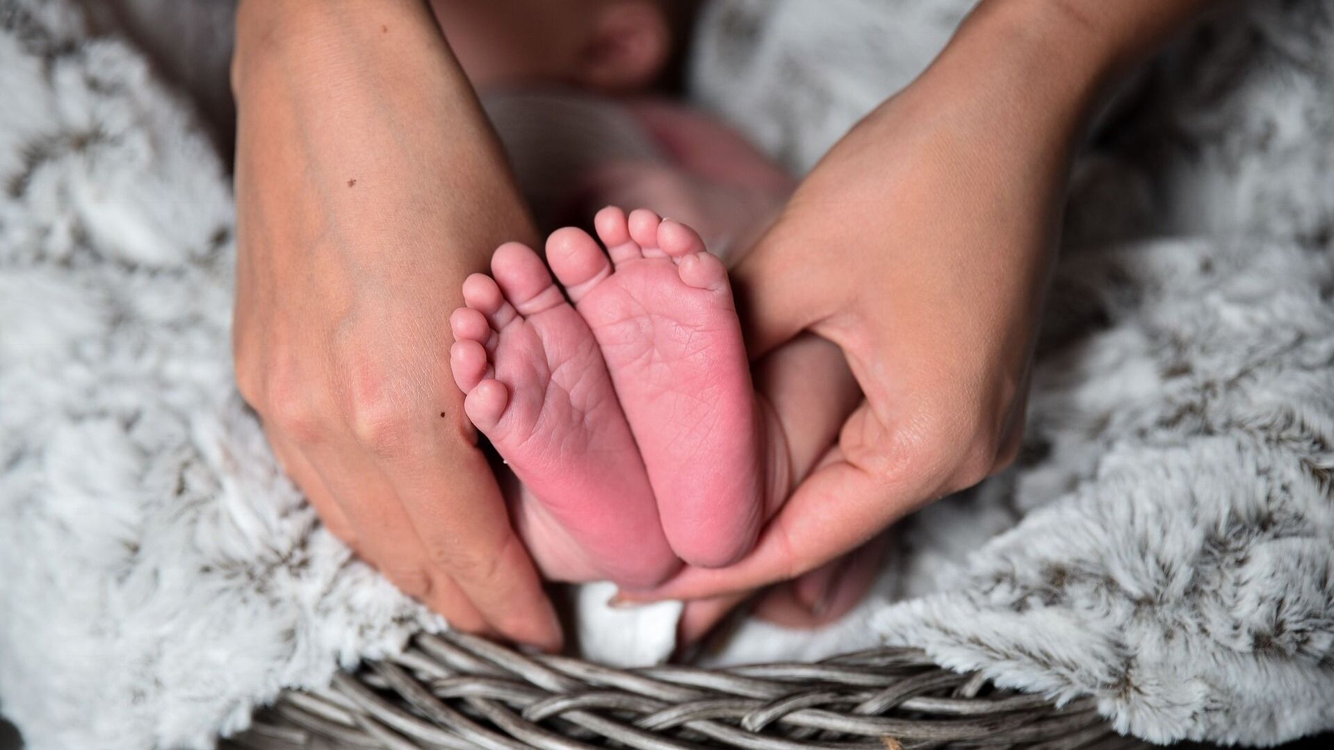 Nahaufnahme von zwei kleinen Babyfüßen, die sanft von zwei Händen gehalten werden, auf einer weichen Decke in einem Korb.