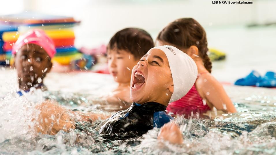 Lachendes Kind mit Schwimmhaube spritzt Wasser im Schwimmbad, umgeben von anderen Kindern in Badebekleidung.
