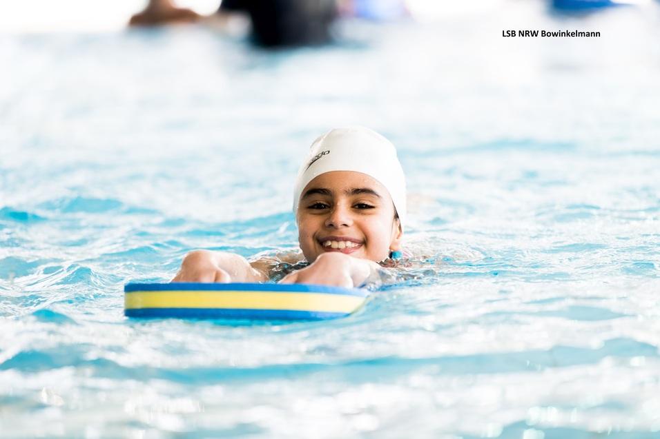 Lächelndes Mädchen mit Badekappe auf einem Schwimmbrett im Wasser eines Hallenbads.
