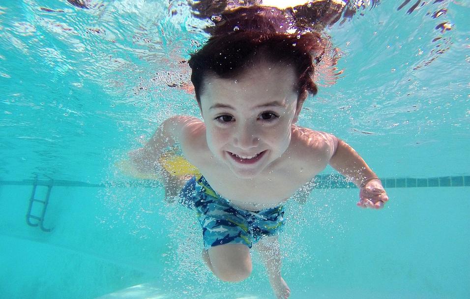 Junge schwimmt unter Wasser mit l&auml;chelndem Gesicht und bunten Schwimmshorts in einem Pool. Blasen entstehen um ihn herum.