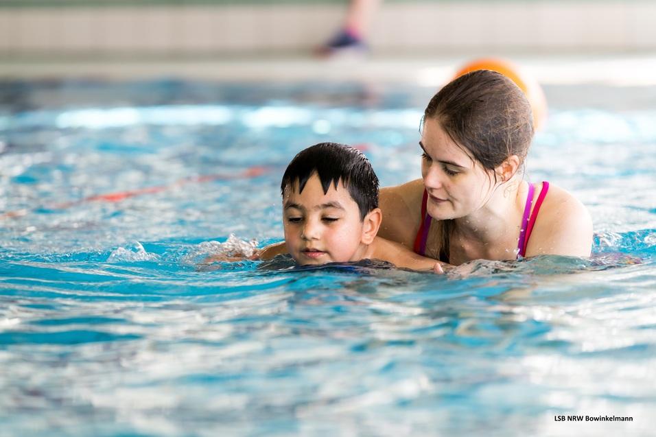 Eine Frau unterstützt ein Kind beim Schwimmen in einem Sportbecken mit blauer Wasseroberfläche.