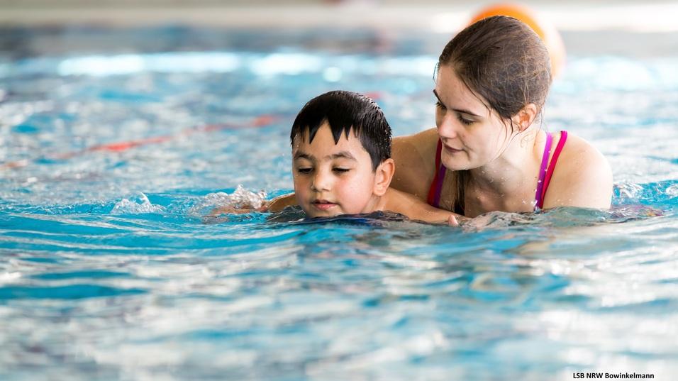 Eine Frau unterstützt ein Kind beim Schwimmen in einem Sportbecken mit blauer Wasseroberfläche.
