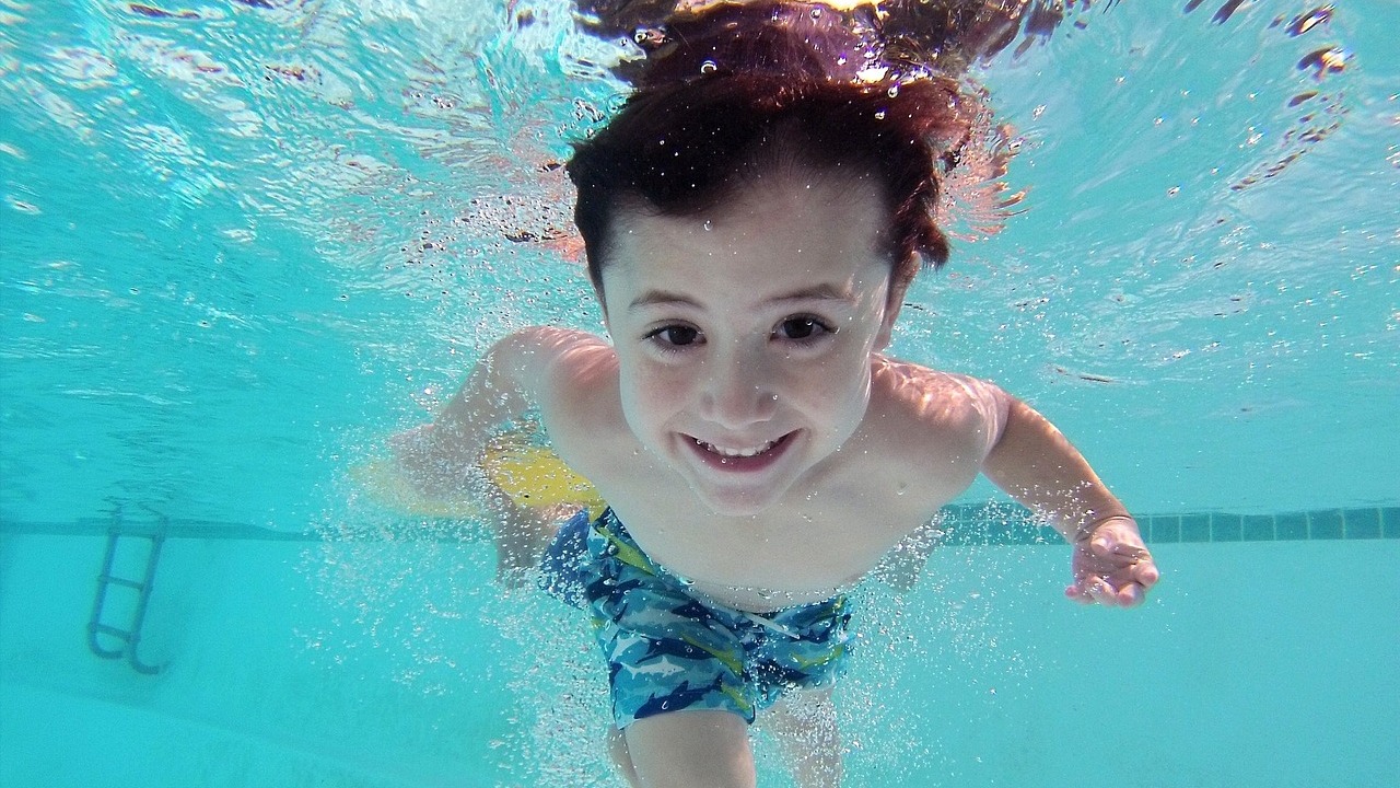 Junge schwimmt lächelnd unter Wasser in einem Pool, umgeben von Blasen und blauem Wasser.