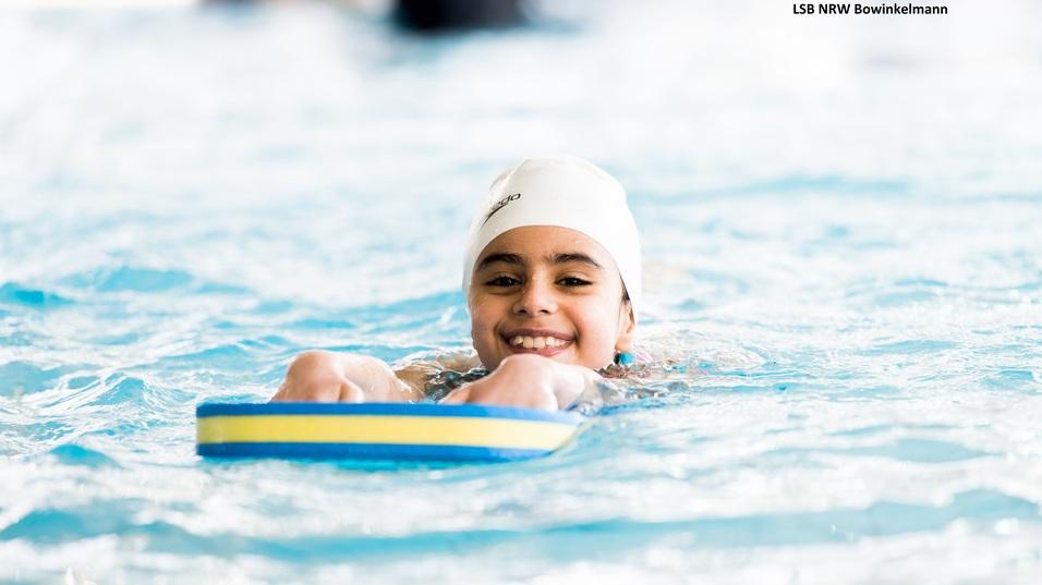 Lächelndes Mädchen mit Badekappe auf einem Schwimmbrett im Wasser eines Hallenbads.