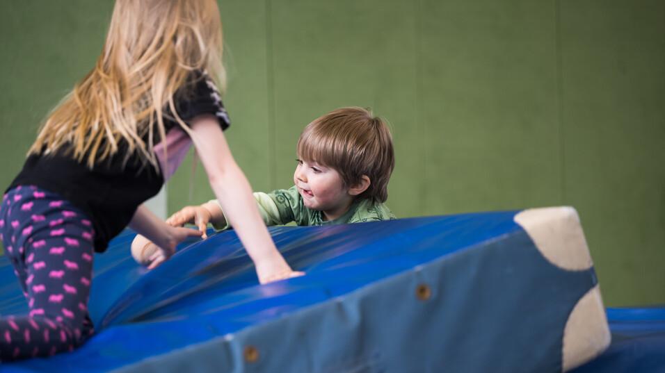 Zwei Kinder spielen auf einer blauen Matratze in einer Sporthalle, das Mädchen zieht den Jungen nach oben.