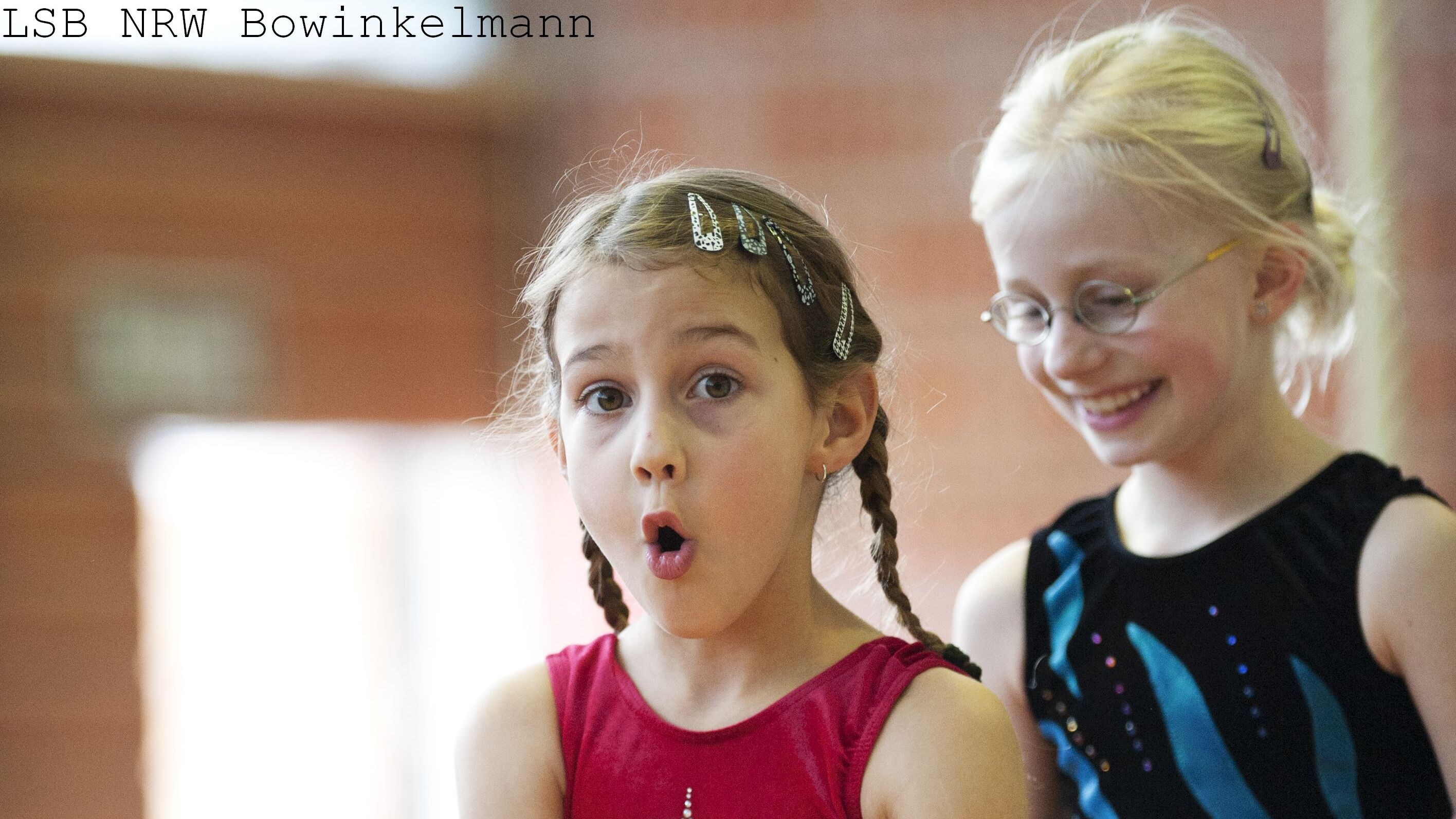 Zwei junge M&auml;dchen in Trainingsanz&uuml;gen stehen in einer Turnhalle, eines zeigt eine &uuml;berraschte Mimik, das andere l&auml;chelt.