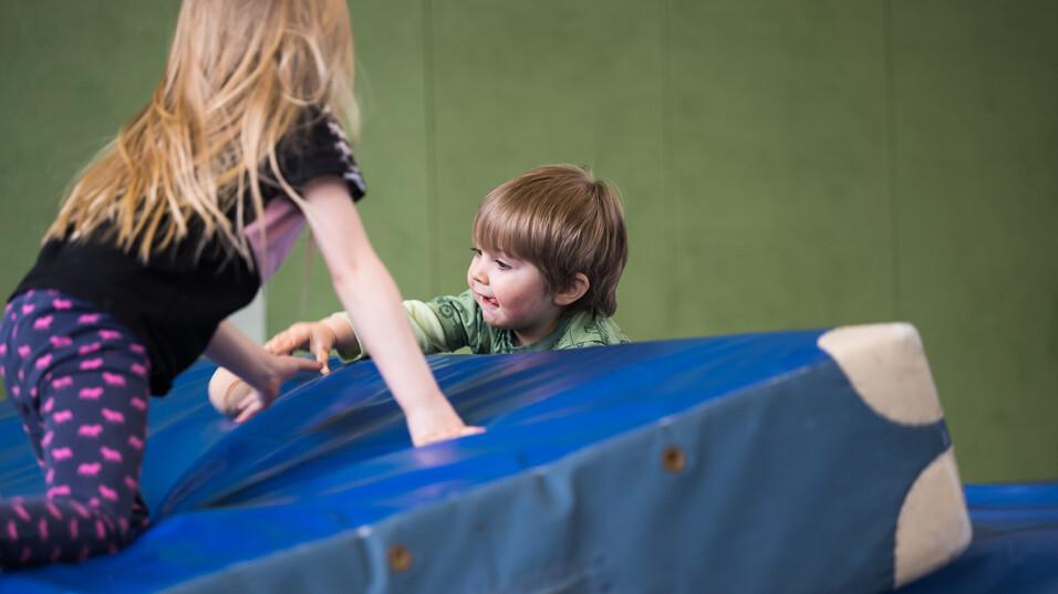 Zwei Kinder spielen auf einer blauen Matratze in einer Sporthalle, das Mädchen zieht den Jungen nach oben.