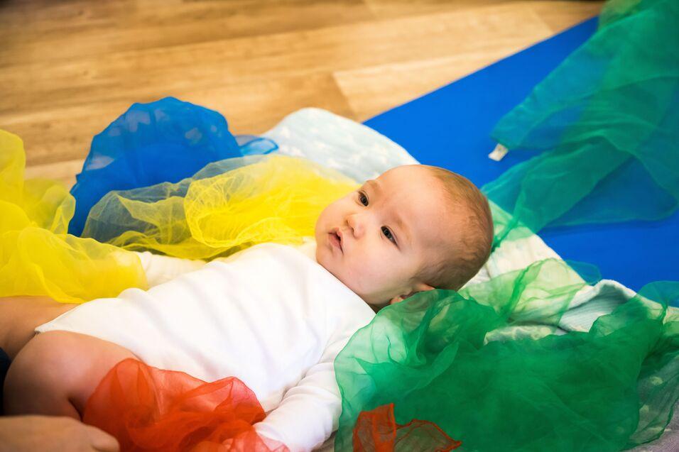 Wachsendes Baby liegt auf einer Matte, umgeben von bunten, transparenten Tüchern in Gelb, Blau, Grün und Rot.