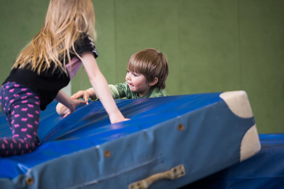 Zwei Kinder spielen auf einer blauen Matratze in einer Sporthalle, das Mädchen zieht den Jungen nach oben.