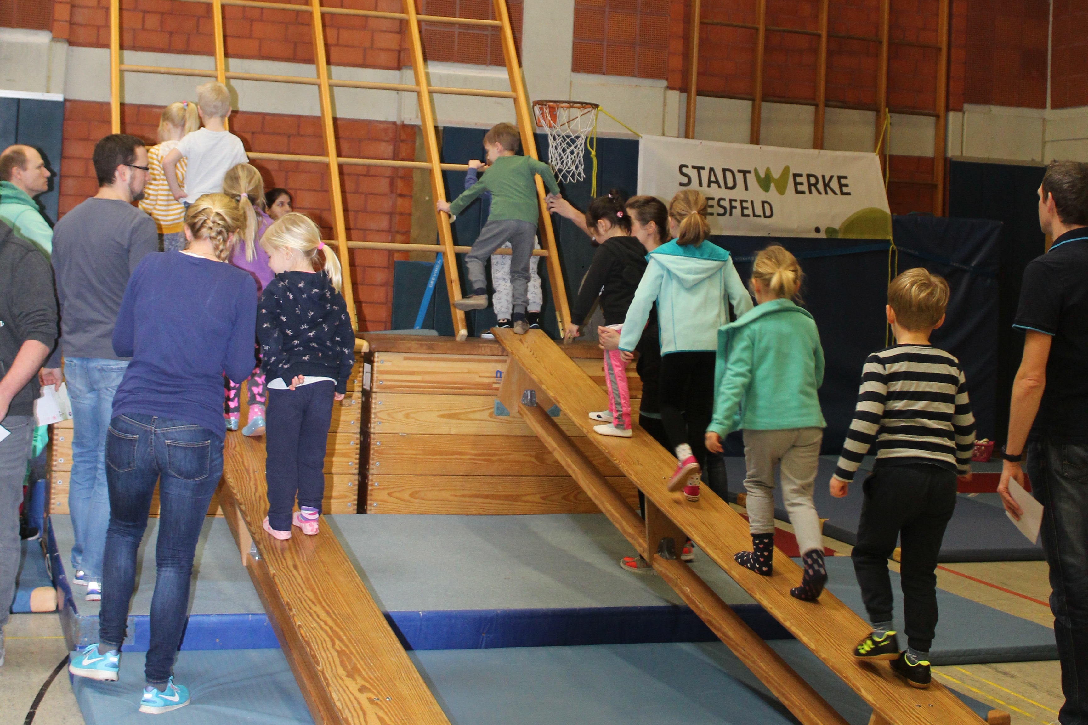 Gruppe von Kindern und Erwachsenen steigt über Holzrampen in einer Sporthalle mit Klettergerüst und Sportgeräten.