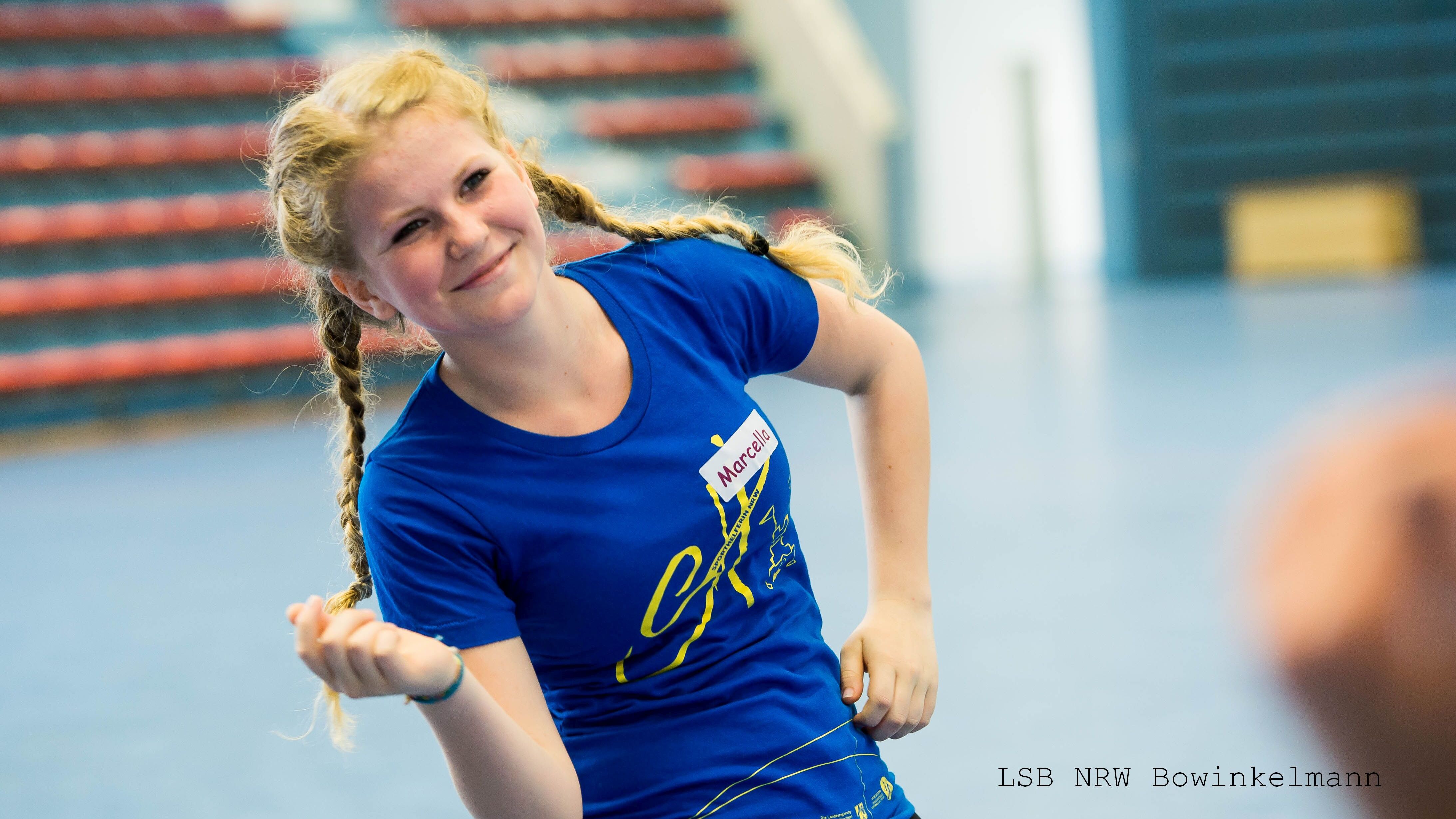 L&auml;chelnde Jugendliche mit Z&ouml;pfen tr&auml;gt ein blaues T-Shirt und bewegt sich in einer Sporthalle.
