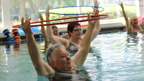 Gruppen von Frauen in einem Schwimmbecken heben Hula-Hoop-Reifen über ihren Köpfen während einer Wasserübungsstunde.