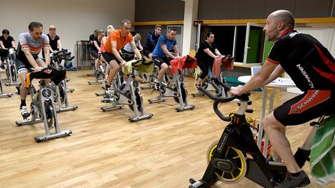 Gruppentraining mit mehreren Personen auf Spinning-Bikes in einem Fitnessraum, ein Trainer gibt Anweisungen.