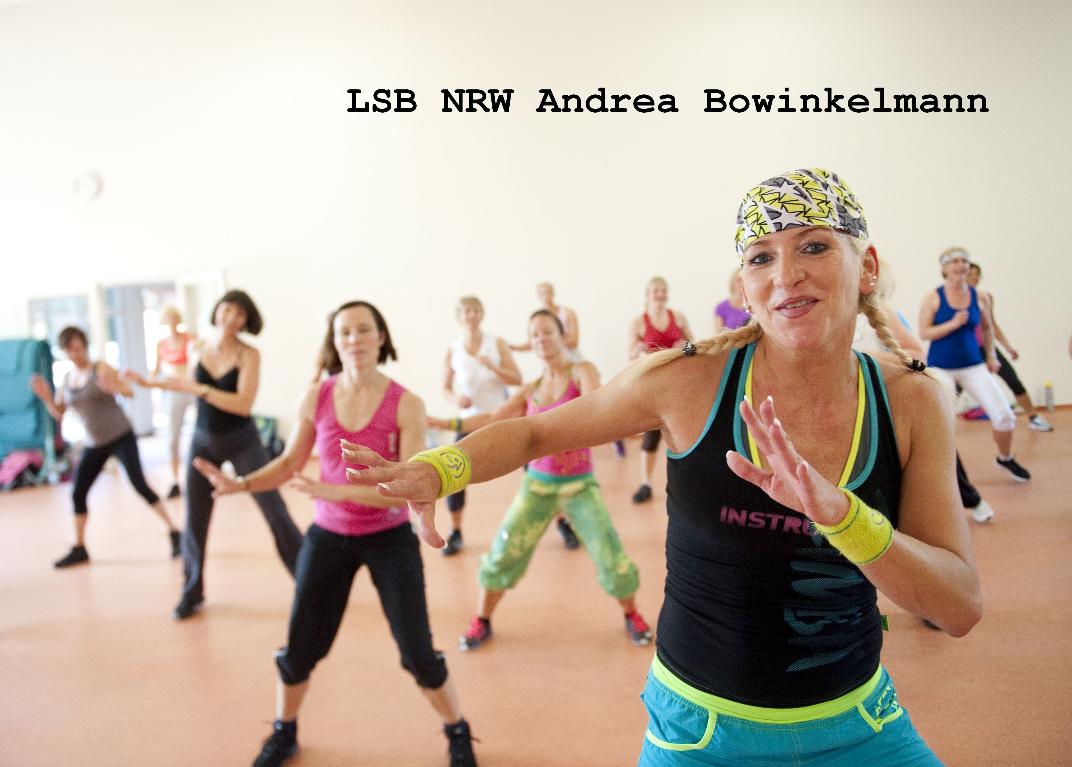 Gruppentraining mit mehreren Frauen in Sportkleidung, die rhythmisch zu Musik tanzen. Im Vordergrund eine Frau mit breitem Lächeln.