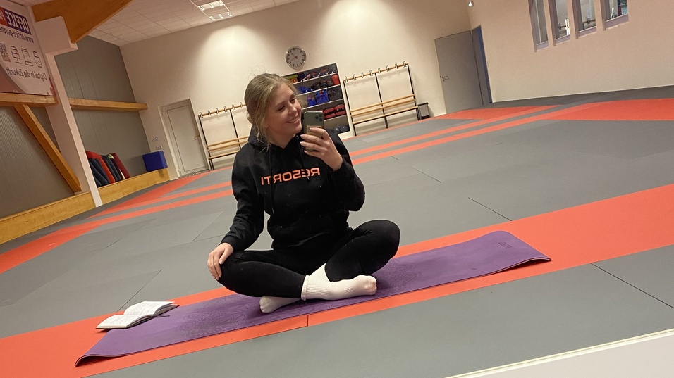 Junge Frau im Sportanzug sitzt auf einer Yogamatte in einem leeren Trainingsraum und lächelt ins Handy.