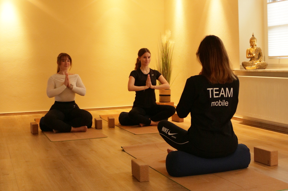 Drei Frauen in Yogastellungen sitzen auf Matten, umgeben von Yoga-Blöcken in einem hellen Raum.