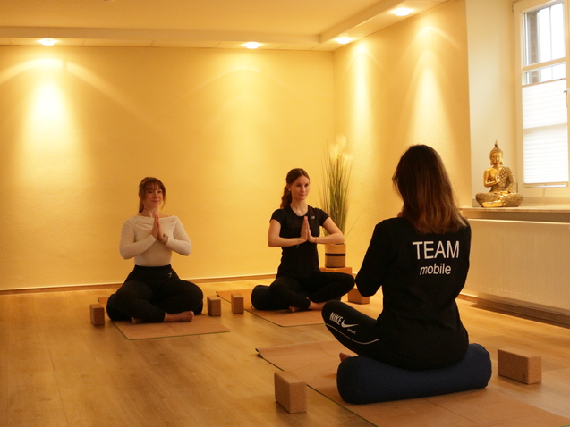 Drei Frauen in Yogastellungen sitzen auf Matten, umgeben von Yoga-Blöcken in einem hellen Raum.
