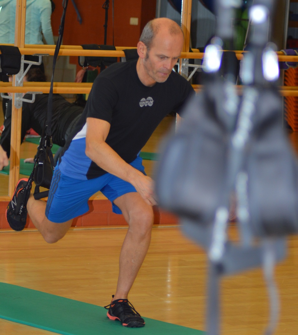 Mann in Sportkleidung führt eine Dehnübung auf einer Trainingsmatte in einem Fitnessraum aus.