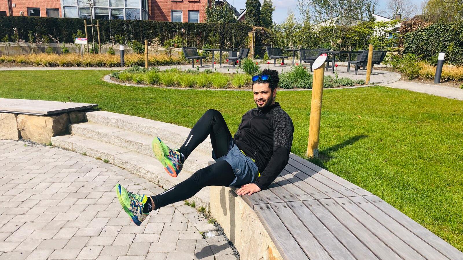 Mann sitzt auf einer Holzbank im Park, seine Beine in Sportkleidung sind hochgehoben, umgeben von Grünflächen.