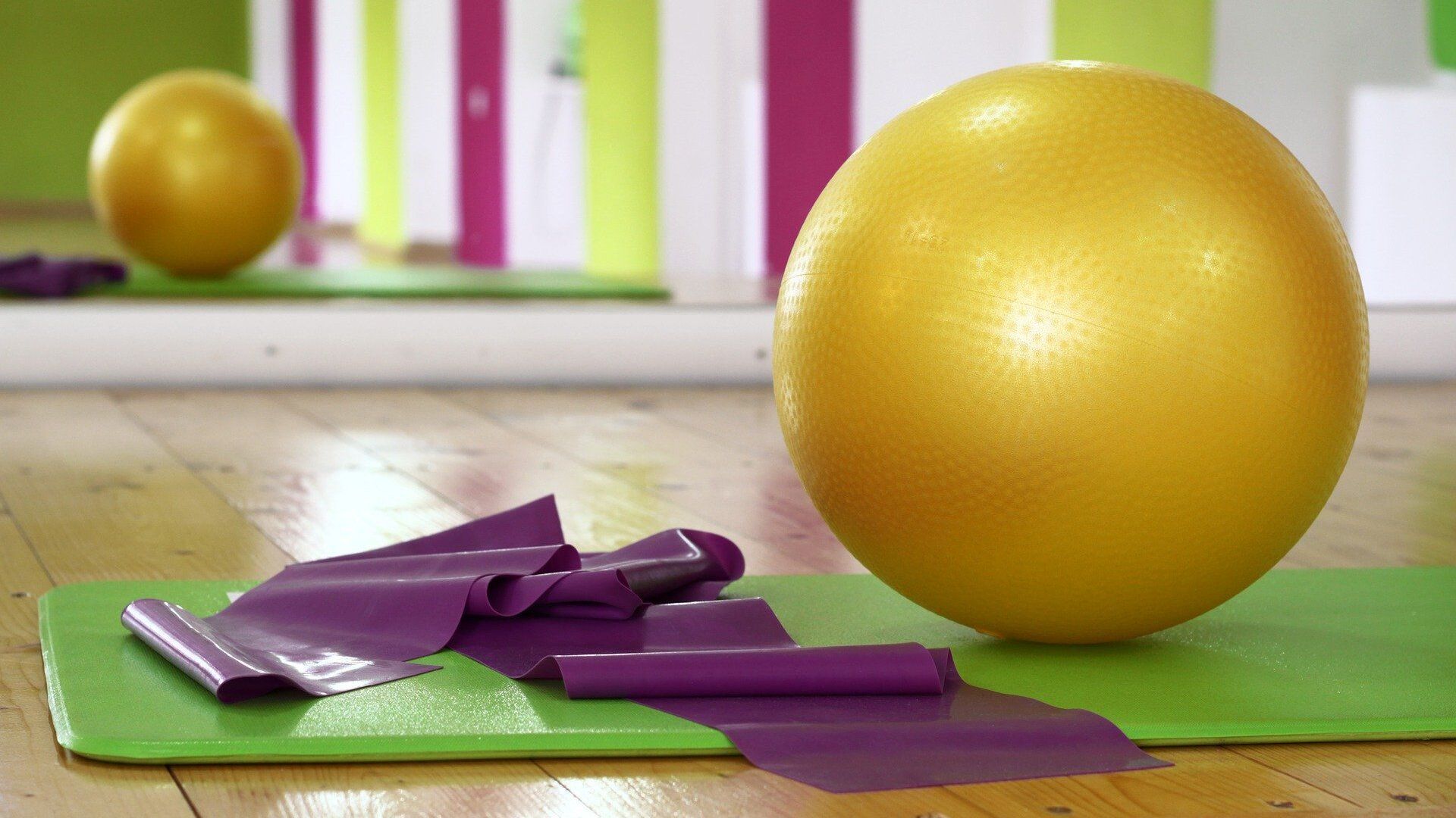 Gelbe Übungsball und purple Widerstandsbänder liegen auf einer grünen Yogamatte in einem hellen Raum mit bunten Wänden.