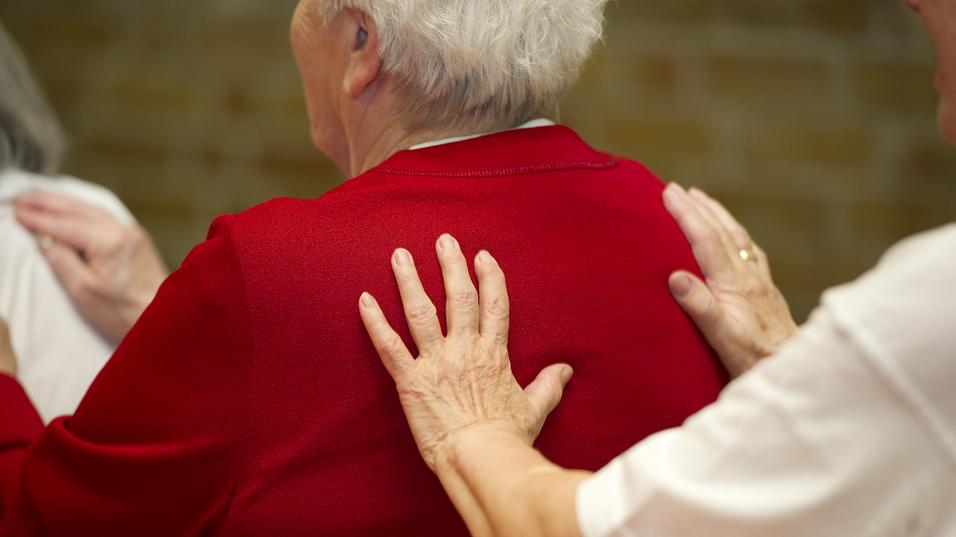 Nahaufnahme von zwei Händen, die sanft die Schultern einer älteren Frau in rotem Pullover berühren.
